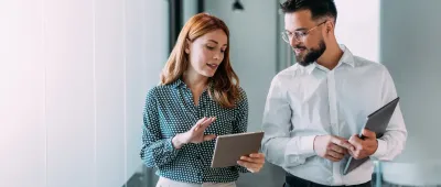 Due professionisti, un uomo e una donna, collaborano analizzando dati su un tablet in un corridoio d'ufficio moderno e luminoso.